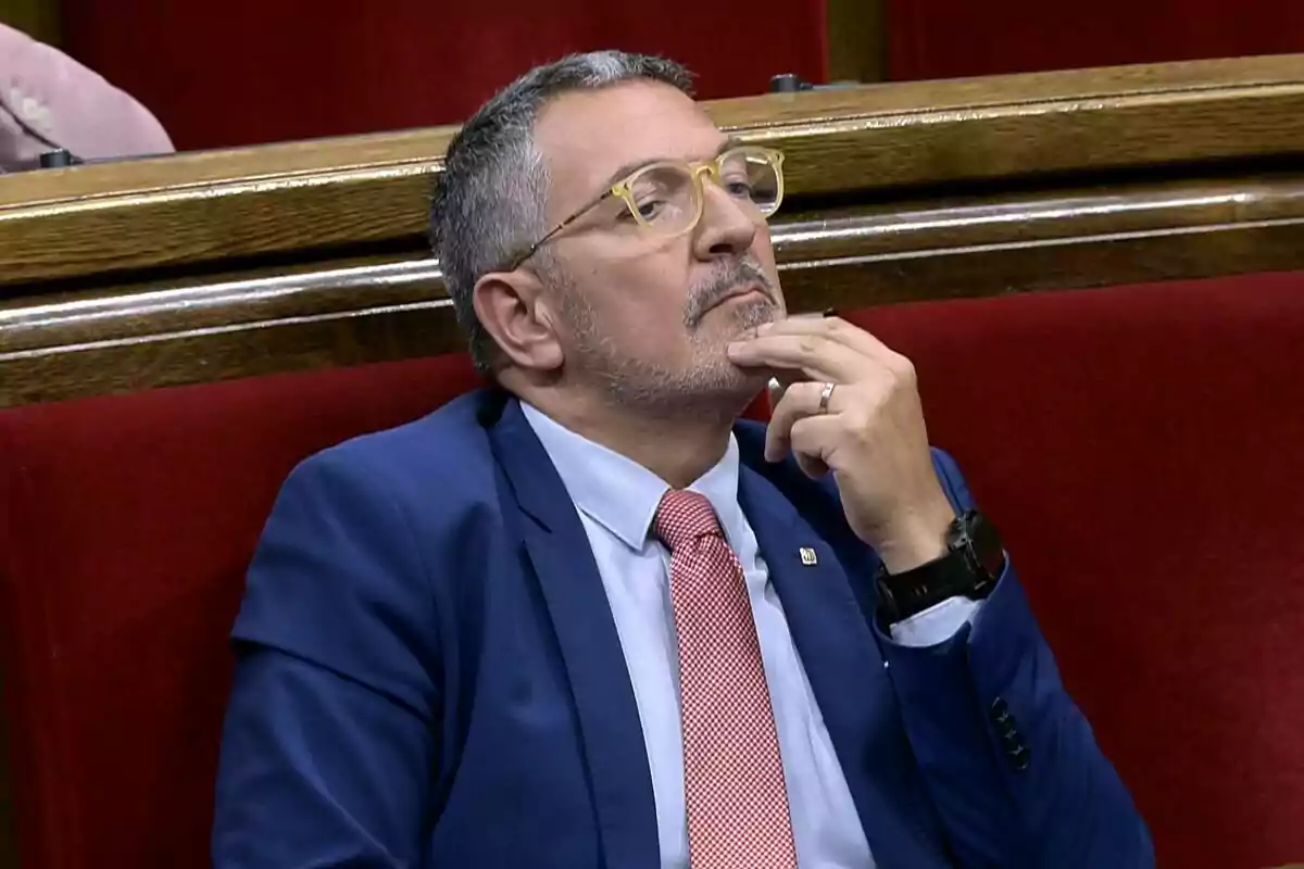 Hombre con gafas y traje azul sentado en un escaño rojo con expresión pensativa y la mano en el mentón