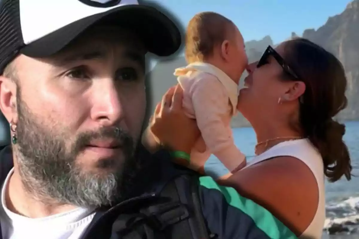 Kiko Rivera con gorra y barba mira pensativo mientras al fondo Anabel Pantoja sostiene y sonríe a un bebé en la playa con montañas detrás