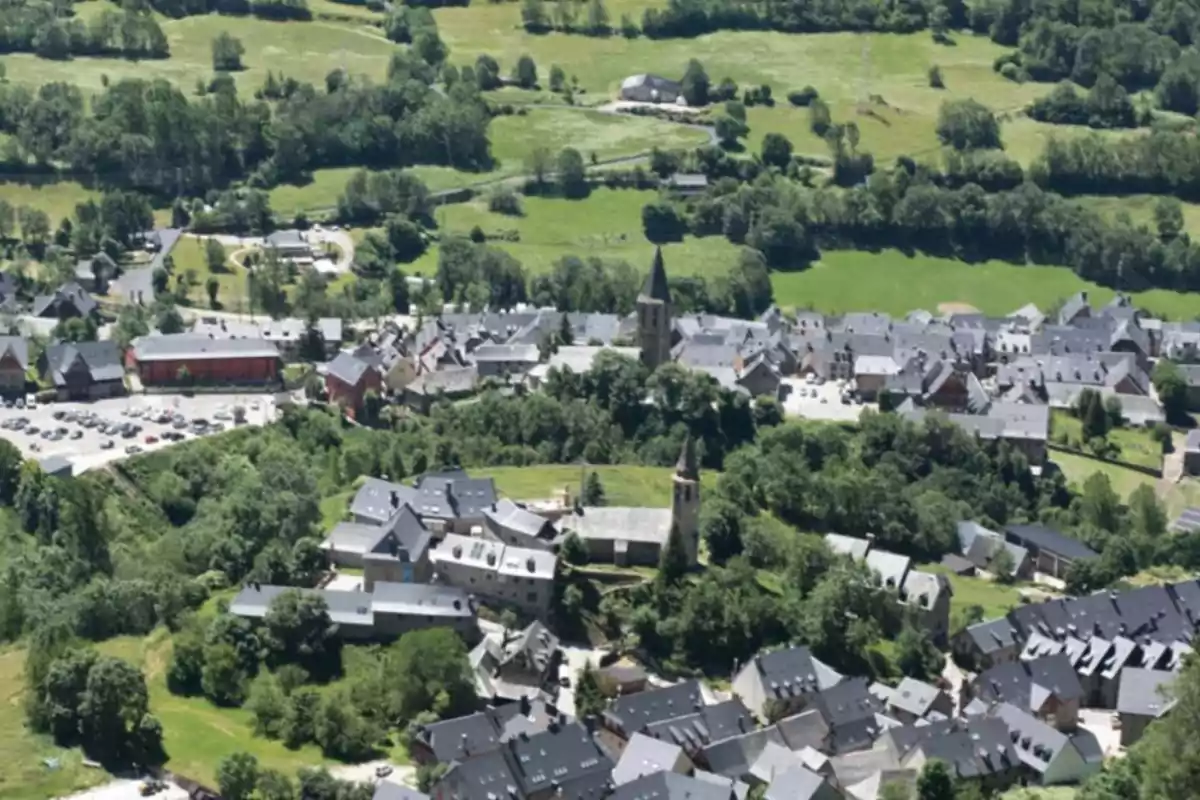 Vista aérea de un pequeño pueblo rodeado de vegetación y campos verdes, con casas de techos oscuros y dos iglesias con campanarios destacados