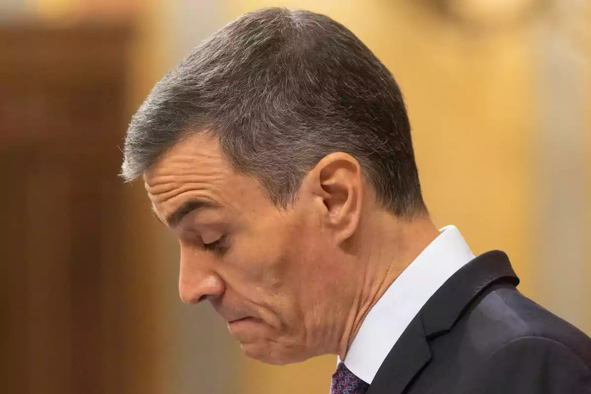 Hombre de cabello canoso con expresión seria mirando hacia abajo en un entorno interior