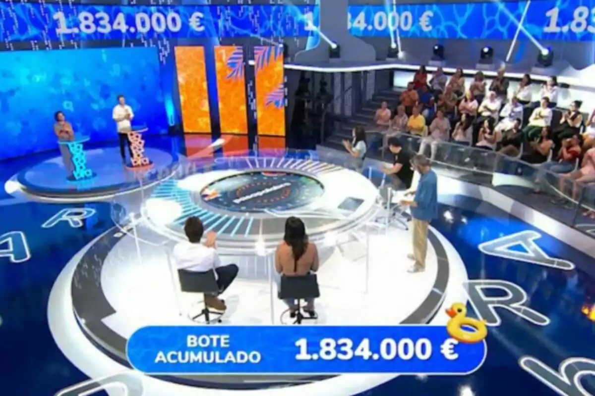 Un plató de concurso televisivo con concursantes, presentador, público y un bote acumulado de un millón ochocientos treinta y cuatro mil euros mostrado en pantalla. Un plató de concurso televisivo con concursantes, presentador, público y un bote acumulado de un millón ochocientos treinta y cuatro mil euros mostrado en pantalla.