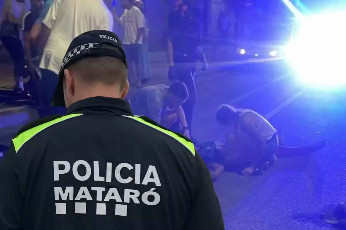 Un agente de la policía de Mataró observa una escena nocturna donde varias personas atienden a alguien en el suelo mientras una luz azul intensa ilumina el lugar