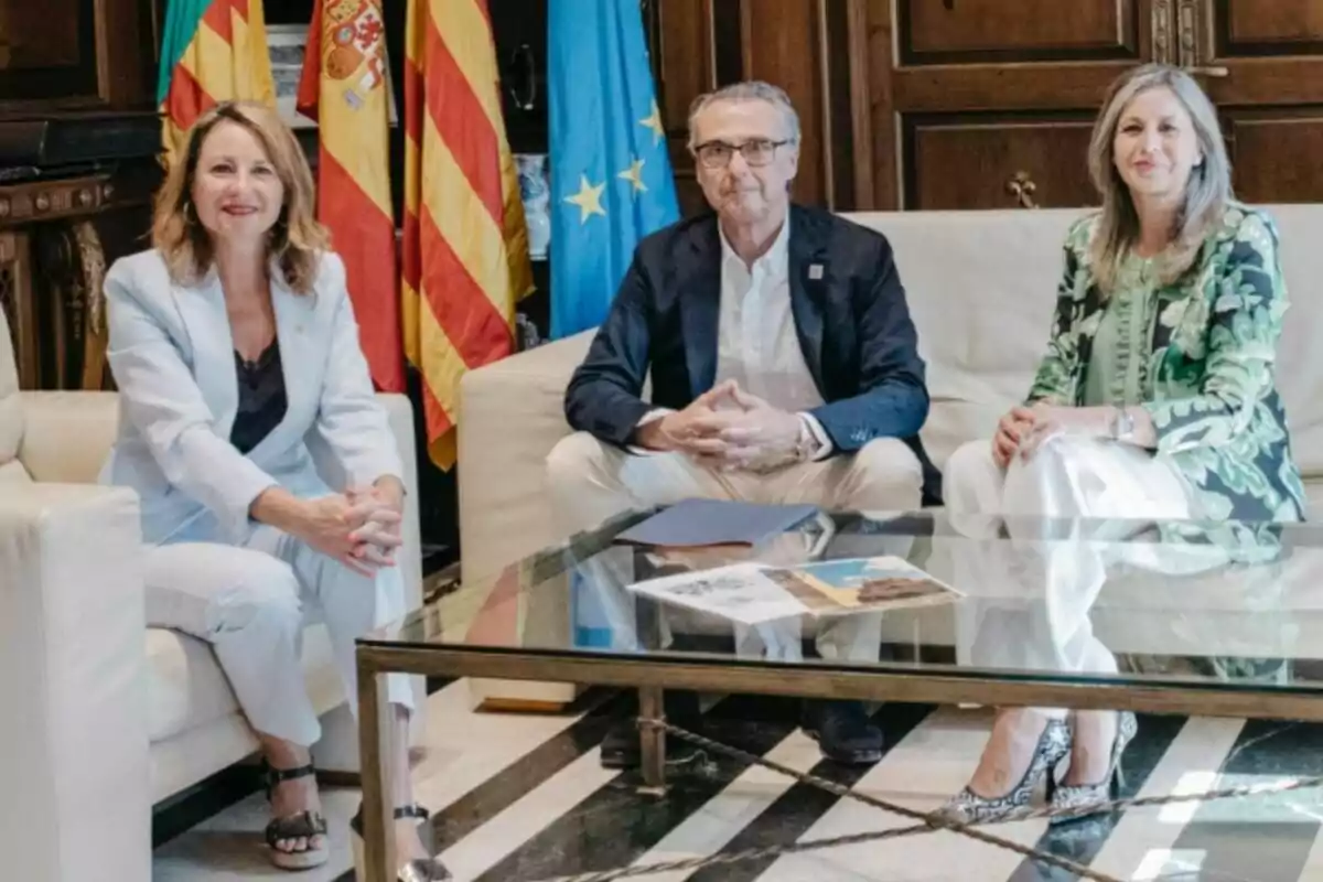 Tres personas sentadas en sofás alrededor de una mesa de cristal en una sala elegante con banderas oficiales al fondo Tres personas sentadas en sofás alrededor de una mesa de cristal en una sala elegante con banderas oficiales al fondo