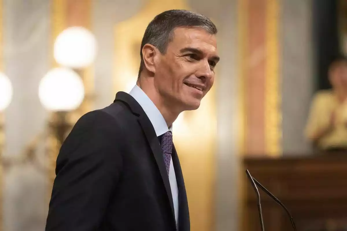 Hombre de cabello corto y canoso con traje oscuro y corbata, sonriendo mientras habla en un micrófono en un entorno elegante y formal Hombre de cabello corto y canoso con traje oscuro y corbata, sonriendo mientras habla en un micrófono en un entorno elegante y formal