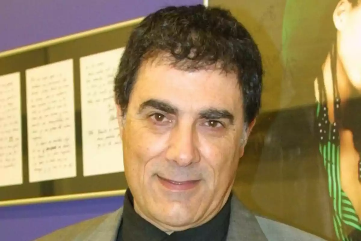 Hombre de cabello oscuro y corto con saco gris y camisa negra sonriendo frente a una pared con documentos enmarcados y una fotografía de fondo