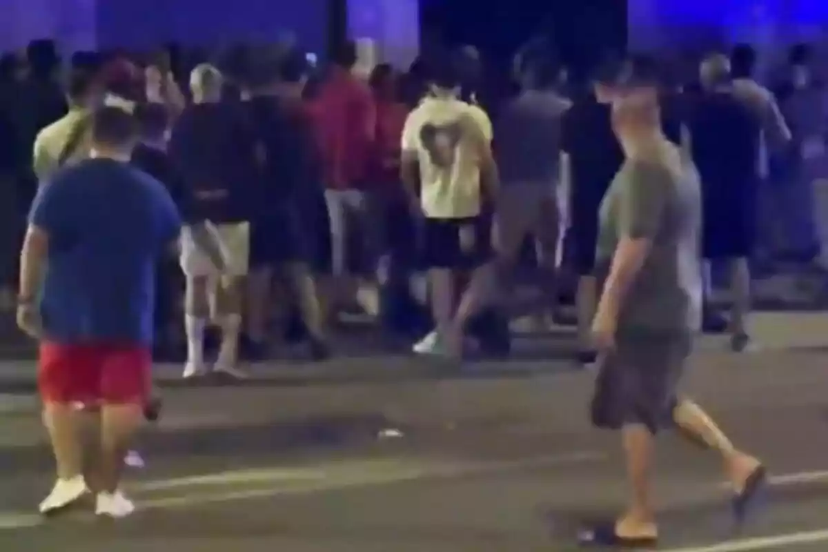 Un grupo de personas reunidas en la calle durante la noche con luces azules al fondo y algunas personas caminando en primer plano.