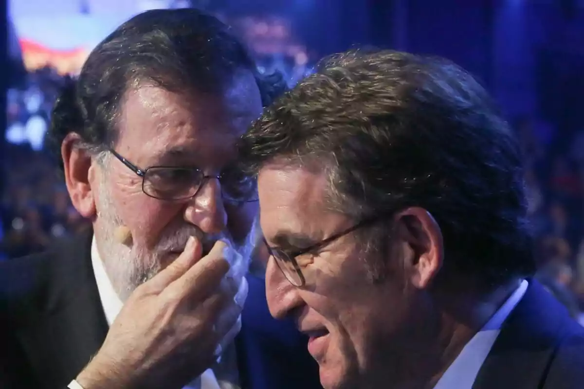 Dos hombres con gafas conversan en un evento, uno de ellos susurra cubriéndose la boca mientras el otro escucha atentamente