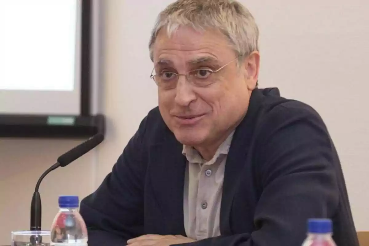 Hombre de cabello canoso y gafas sentado frente a un micrófono en una mesa con botellas de agua