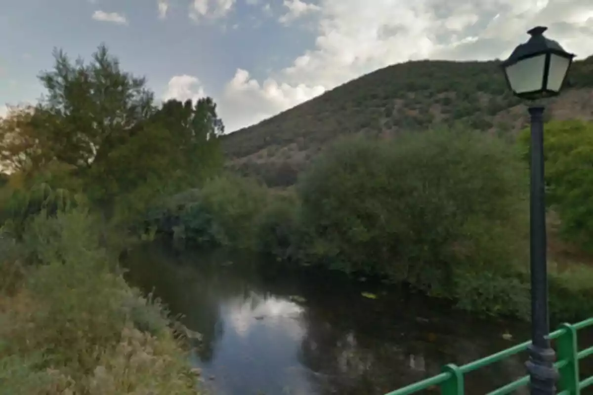 Río Bernesga rodeado de vegetación con una colina al fondo y una farola junto a una barandilla verde en primer plano. Río Bernesga rodeado de vegetación con una colina al fondo y una farola junto a una barandilla verde en primer plano.