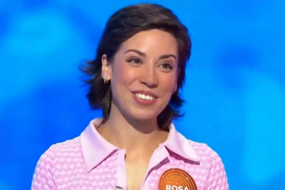 Rosa Rodríguez, concursante de 'Pasapalabra', sonríe mientras participa en este programa de televisión, lleva una camisa rosa con cuello y un pin naranja con el nombre Rosa sobre un fondo azul. Rosa Rodríguez, concursante de 'Pasapalabra', sonríe mientras participa en este programa de televisión, lleva una camisa rosa con cuello y un pin naranja con el nombre Rosa sobre un fondo azul.