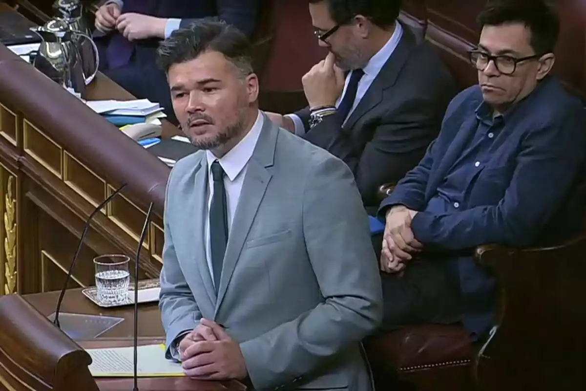 Un hombre con traje gris y corbata verde habla en un atril en una sala parlamentaria mientras otras personas lo observan sentadas detrás de él