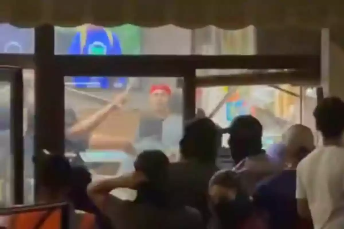 Varias personas observan una pelea al interior de un local mientras un hombre con gorra roja está en el centro de la escena. Varias personas observan una pelea al interior de un local mientras un hombre con gorra roja está en el centro de la escena.