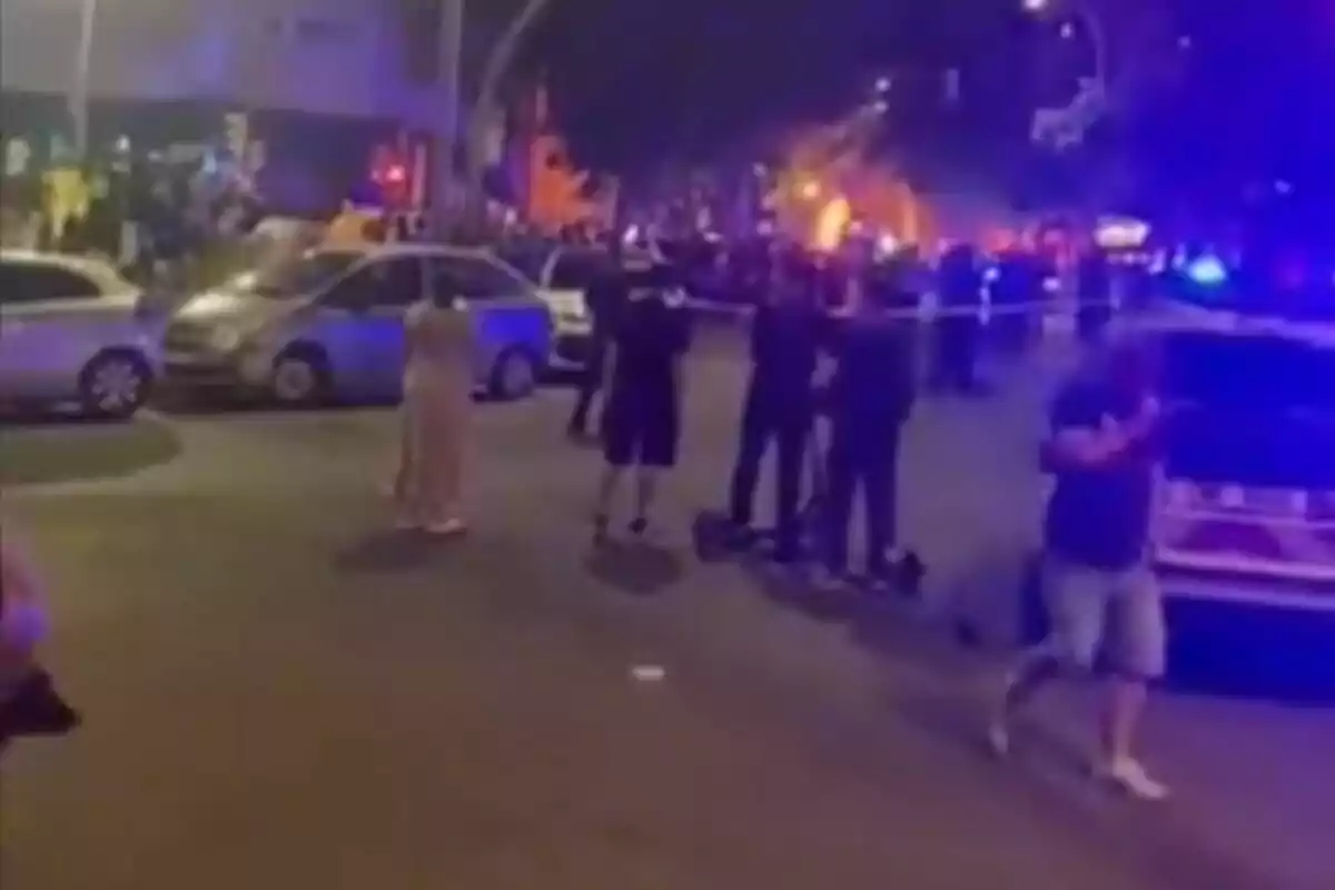Varias personas observan una escena acordonada por la policía en una calle de noche con luces de emergencia y fuego al fondo.