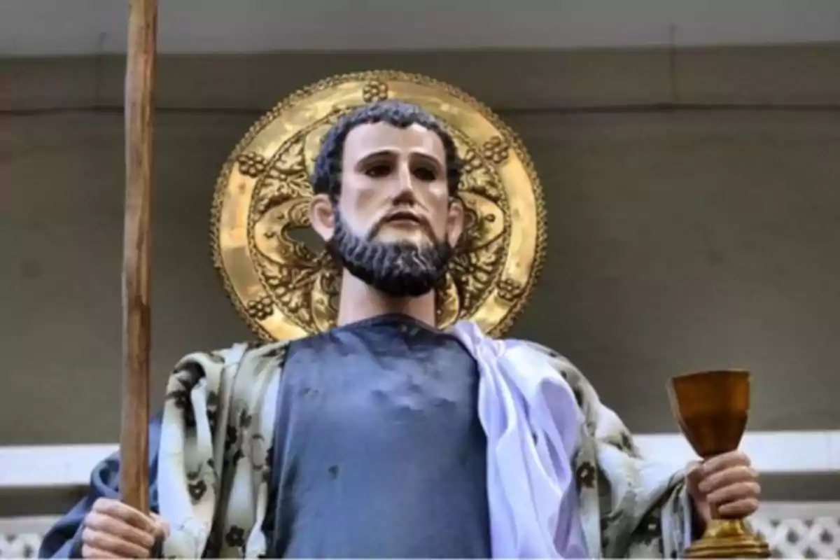 Escultura religiosa de un hombre con barba que sostiene un bastón en una mano y una copa dorada en la otra, con un halo dorado detrás de su cabeza Escultura religiosa de un hombre con barba que sostiene un bastón en una mano y una copa dorada en la otra, con un halo dorado detrás de su cabeza