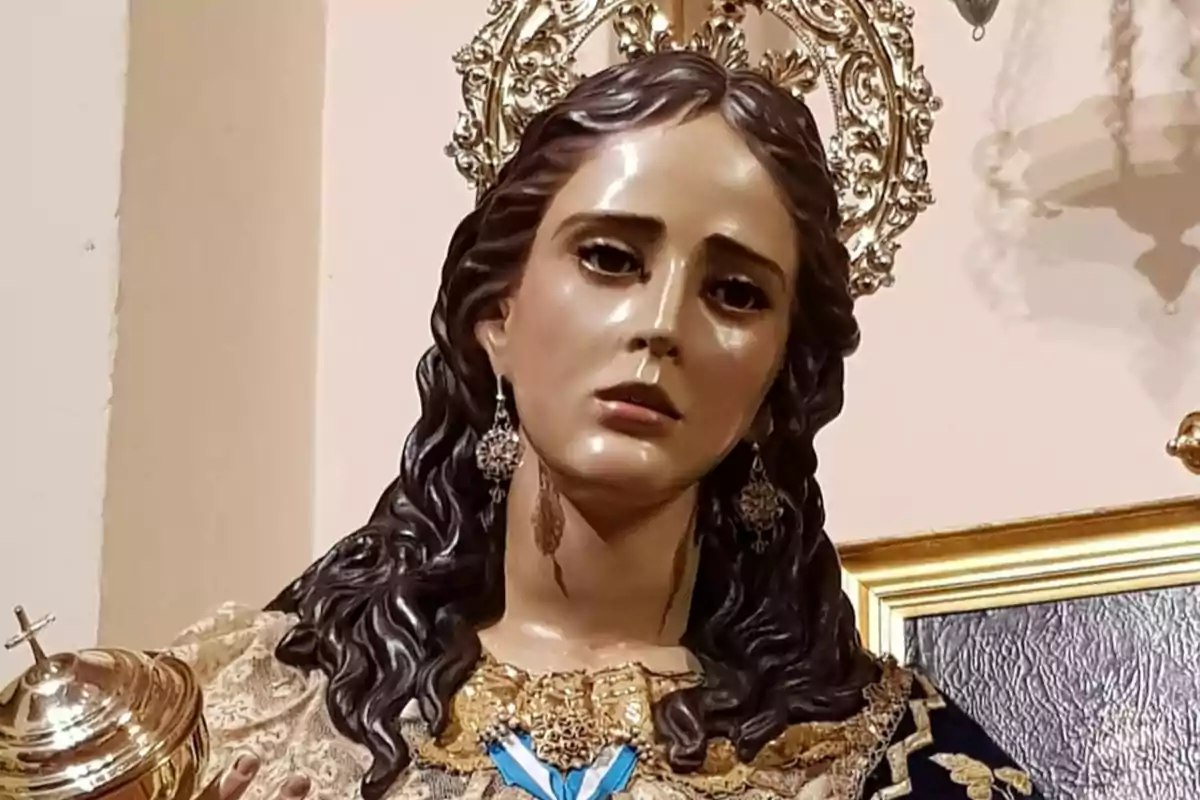 Imagen de una escultura religiosa de una virgen con expresión solemne, cabello oscuro y rizado, vestida con ropajes decorados y una corona dorada detrás de la cabeza