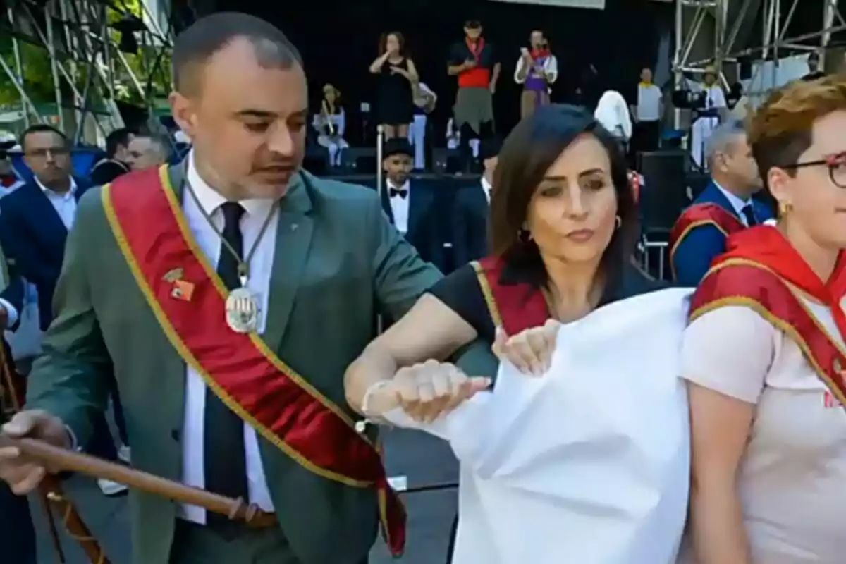 Personas vestidas con bandas rojas y medallas participan en un evento al aire libre, sosteniendo una tela blanca frente a un escenario donde hay más personas