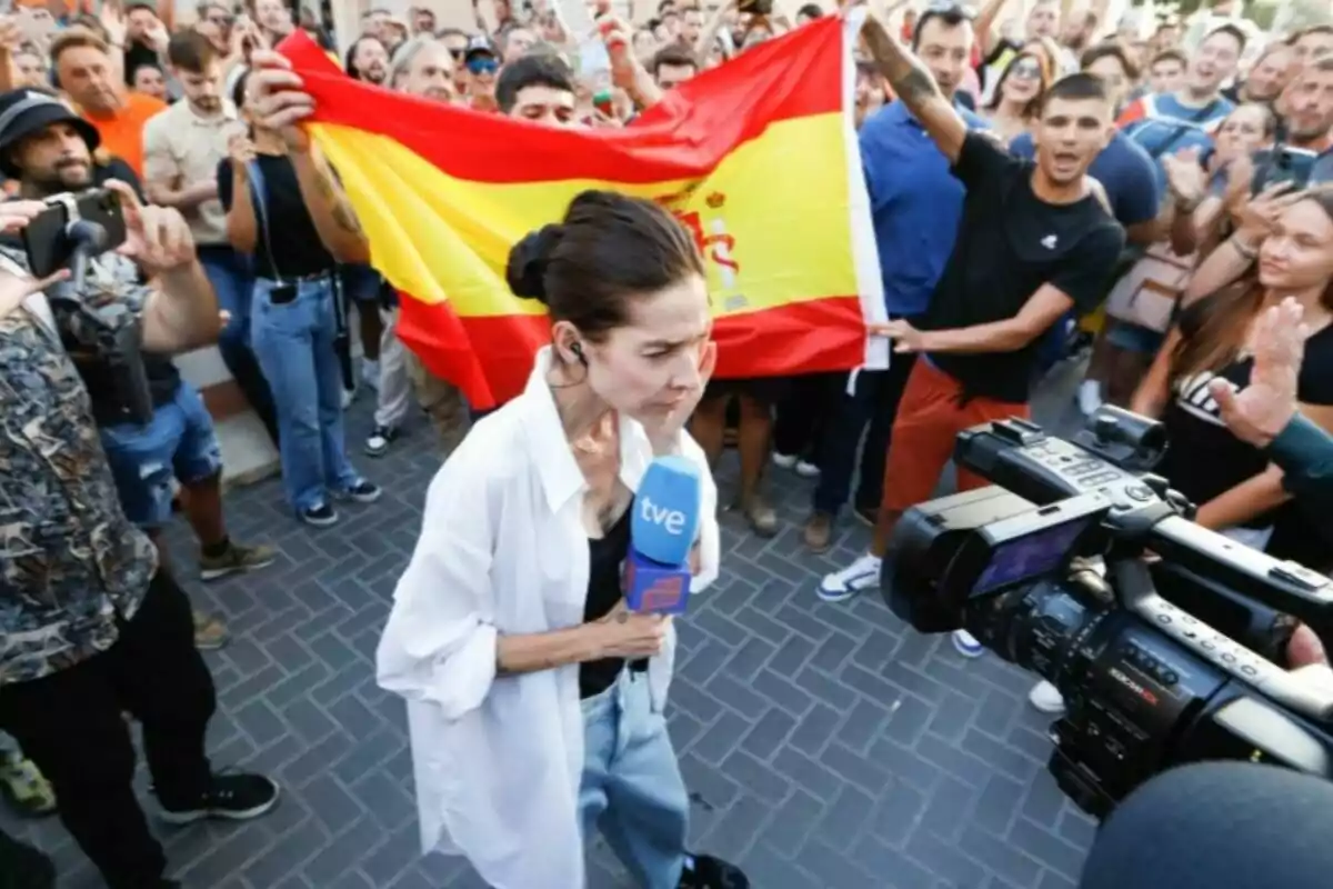 Una reportera sostiene un micrófono mientras es grabada por una cámara rodeada de una multitud que sostiene una bandera de España. Una reportera sostiene un micrófono mientras es grabada por una cámara rodeada de una multitud que sostiene una bandera de España.