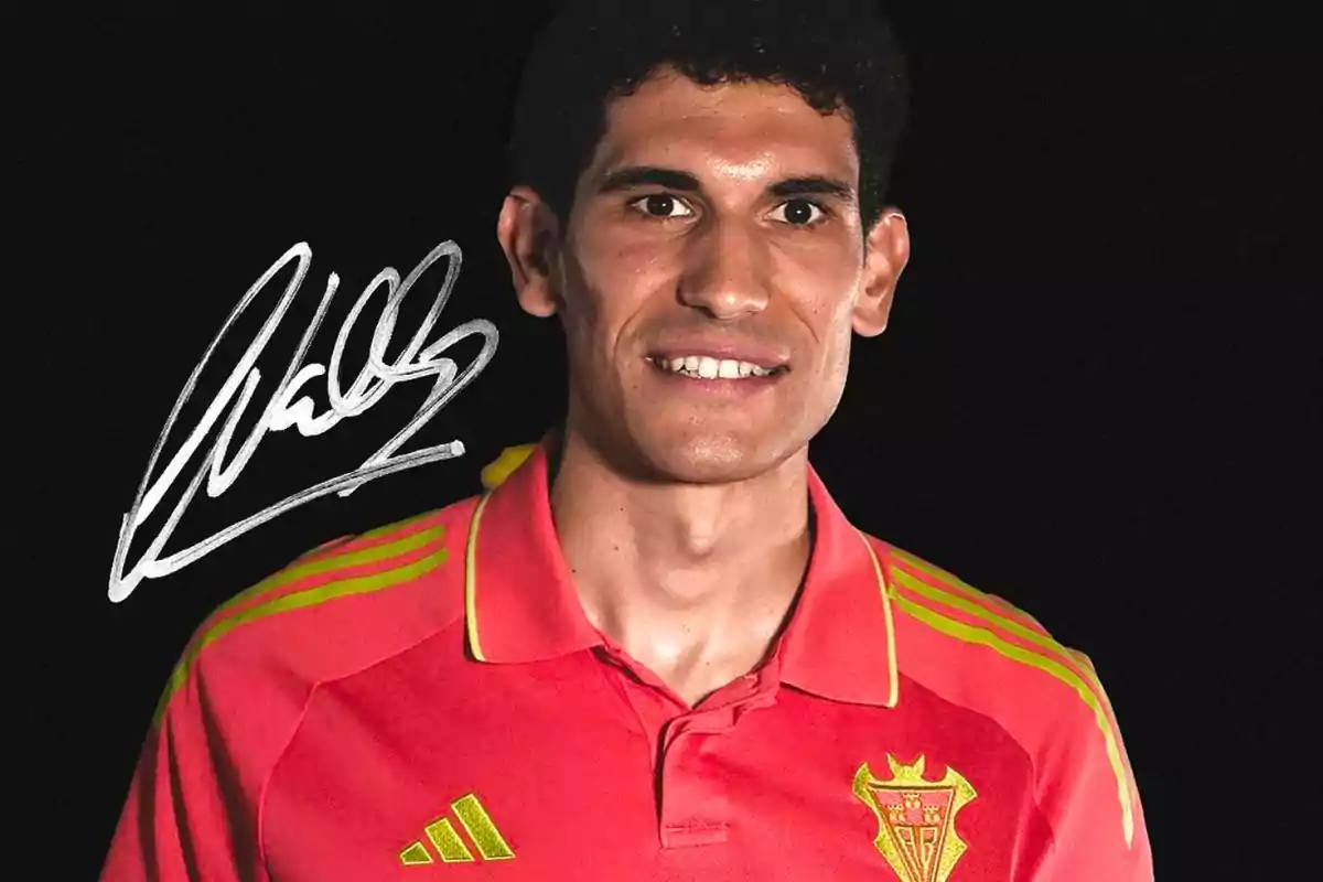 Jugador de fútbol con camiseta roja y detalles amarillos posando frente a un fondo negro con una firma blanca a la izquierda Jugador de fútbol con camiseta roja y detalles amarillos posando frente a un fondo negro con una firma blanca a la izquierda