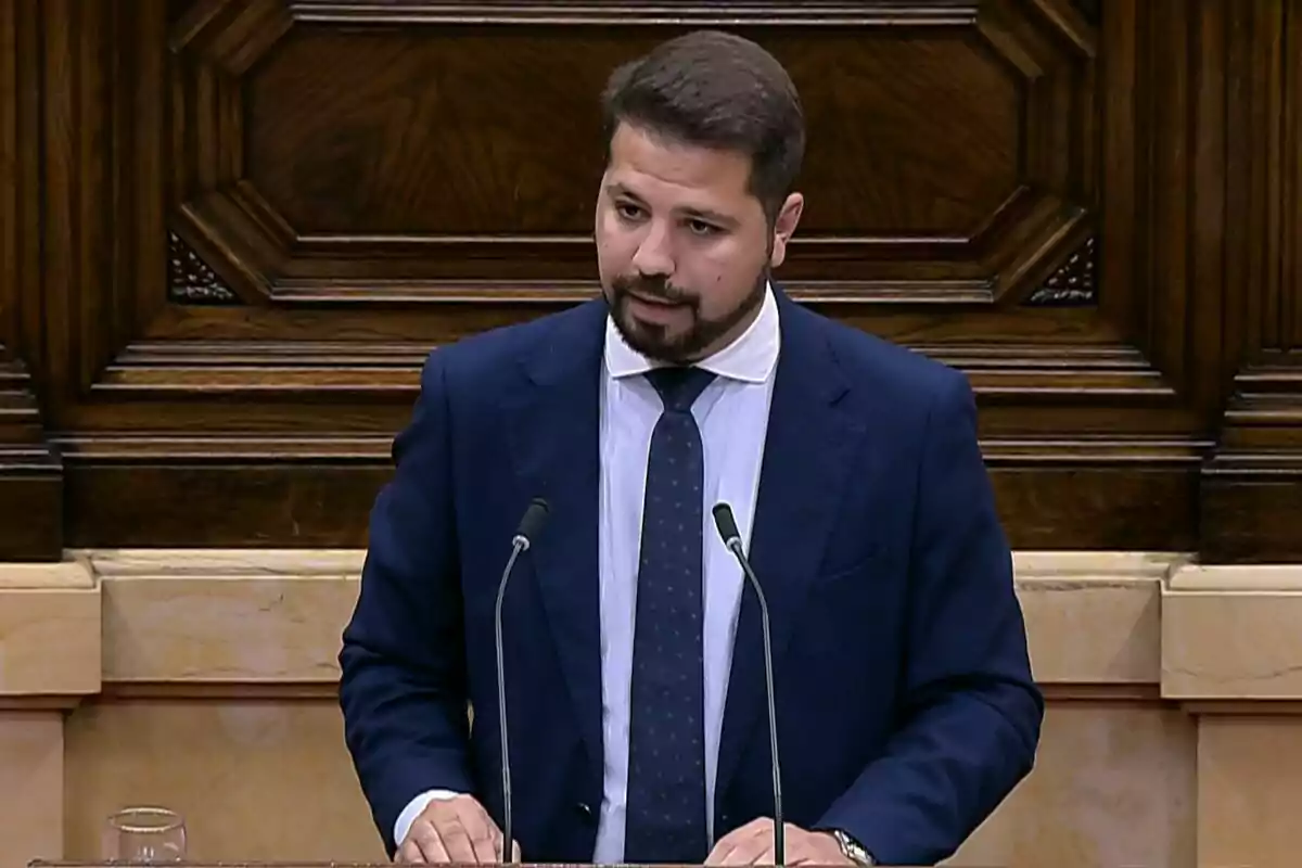 Hombre con barba y traje azul hablando en un atril con dos micrófonos en un entorno de madera elegante Hombre con barba y traje azul hablando en un atril con dos micrófonos en un entorno de madera elegante