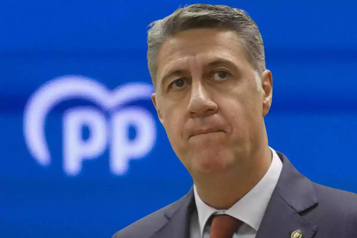 Hombre de traje con expresión seria frente a un fondo azul con el logotipo del Partido Popular desenfocado