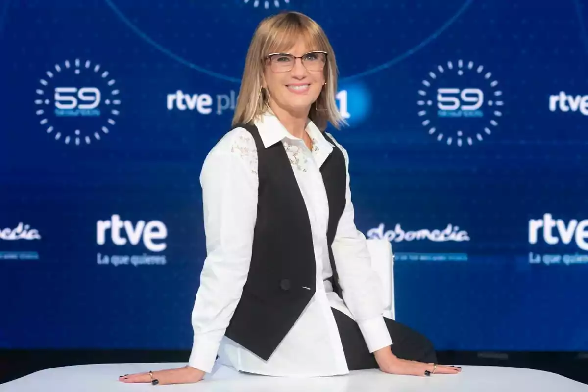 Gemma Nierga con gafas y cabello rubio lacio, vestida con una camisa blanca y un chaleco negro, está sentada en una mesa frente a un fondo azul con logotipos de RTVE y 