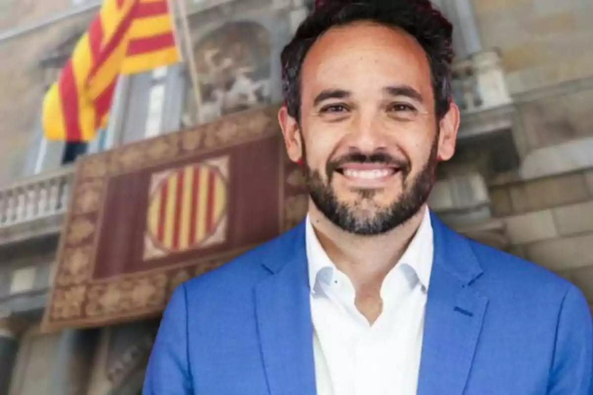 Un hombre con barba y cabello oscuro, vestido con un traje azul y camisa blanca, sonríe frente a un edificio con una bandera catalana ondeando en el fondo.