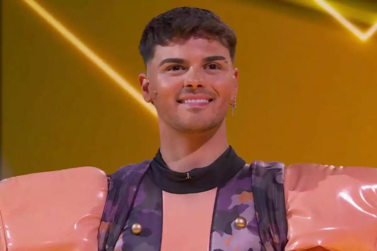 Abraham Mateo sonriente con un atuendo colorido y hombreras grandes en un fondo amarillo en Mask Singer.