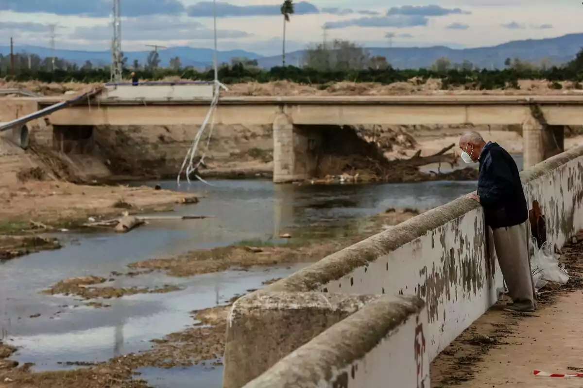 Un hombre con mascarilla observa un río desde un puente en un paisaje rural con montañas al fondo.