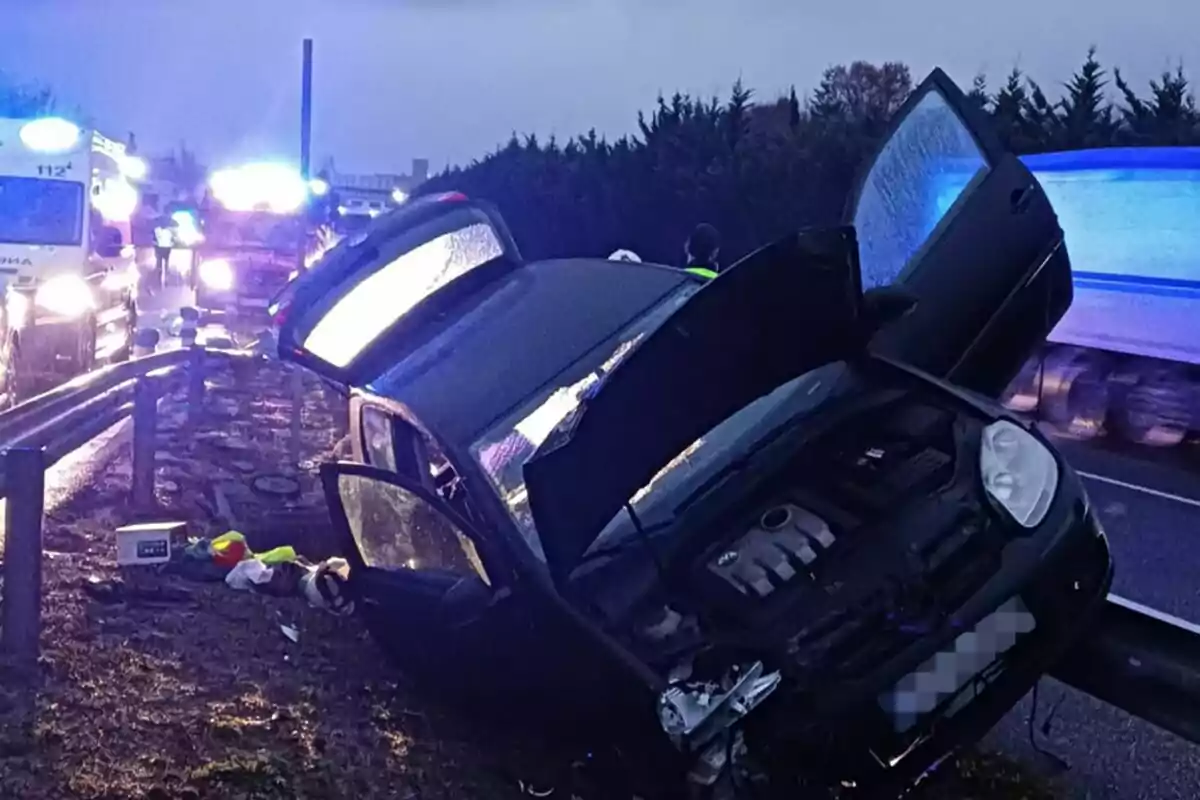 Un coche accidentado en el borde de una carretera con luces de emergencia de vehículos de rescate en el fondo.