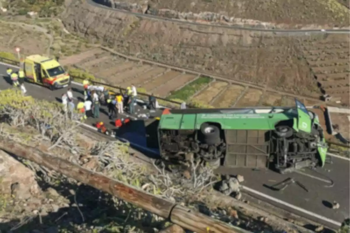 Un autobús verde volcado en una carretera con una ambulancia y varias personas alrededor en un entorno montañoso.