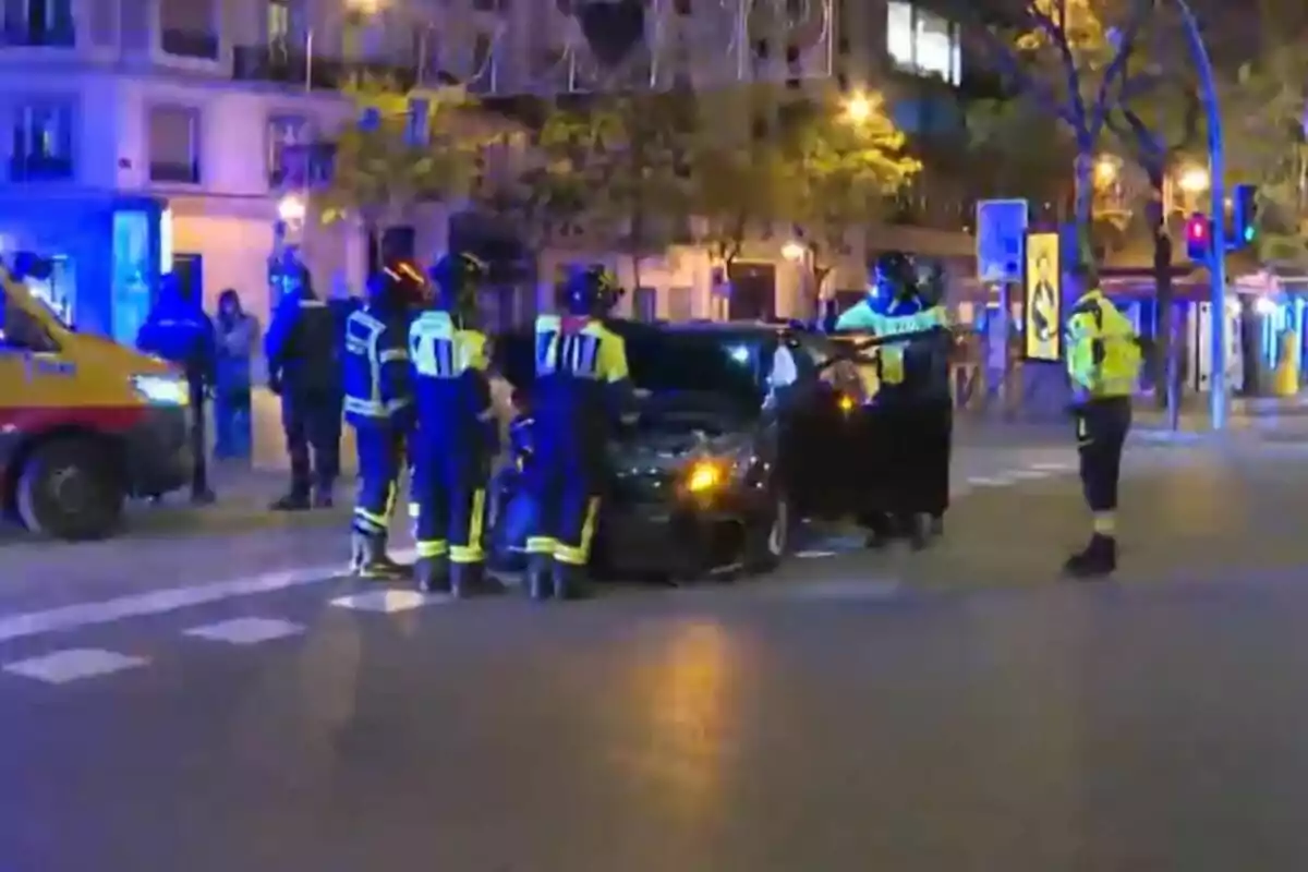 Un grupo de bomberos y policías rodea un coche en una intersección iluminada por luces azules en una ciudad por la noche.