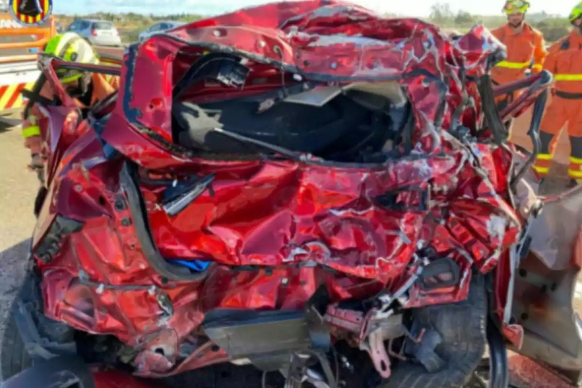 Coche rojo gravemente dañado tras un accidente con bomberos trabajando en el lugar.