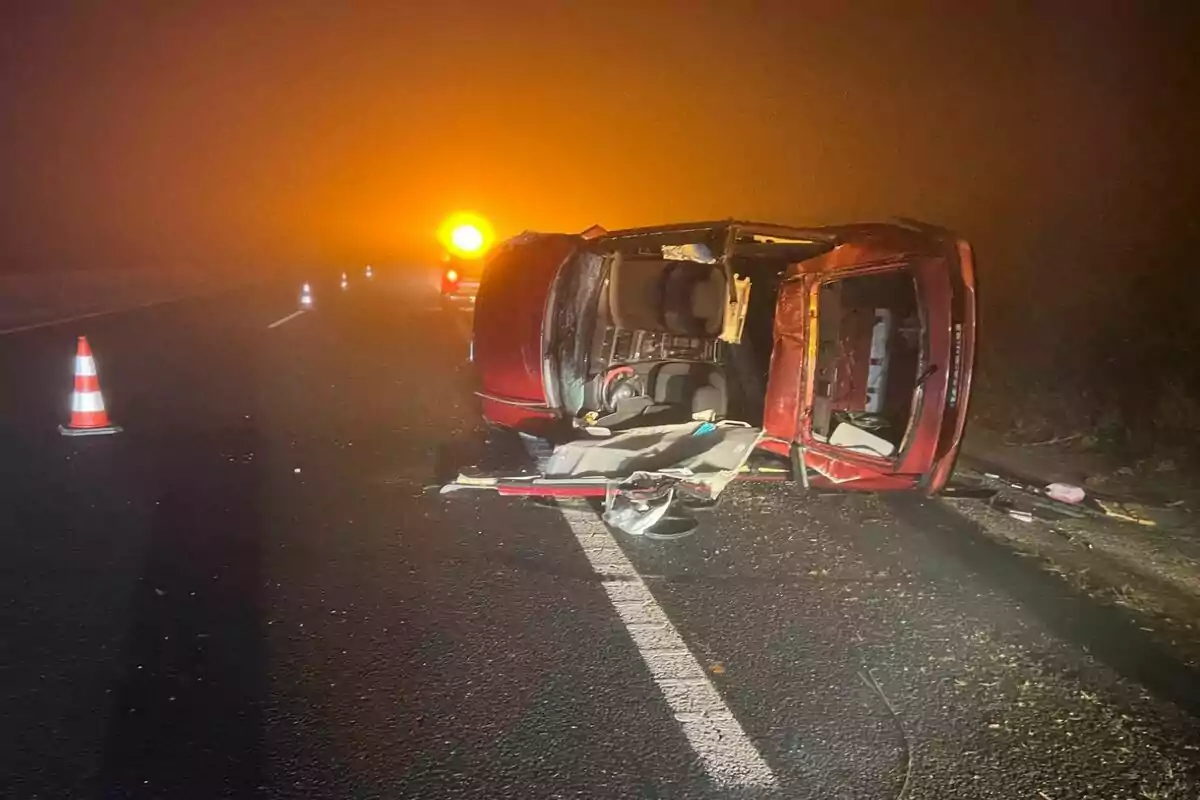 Un coche volcado en una carretera con conos de tráfico y luces de advertencia en una noche con niebla.