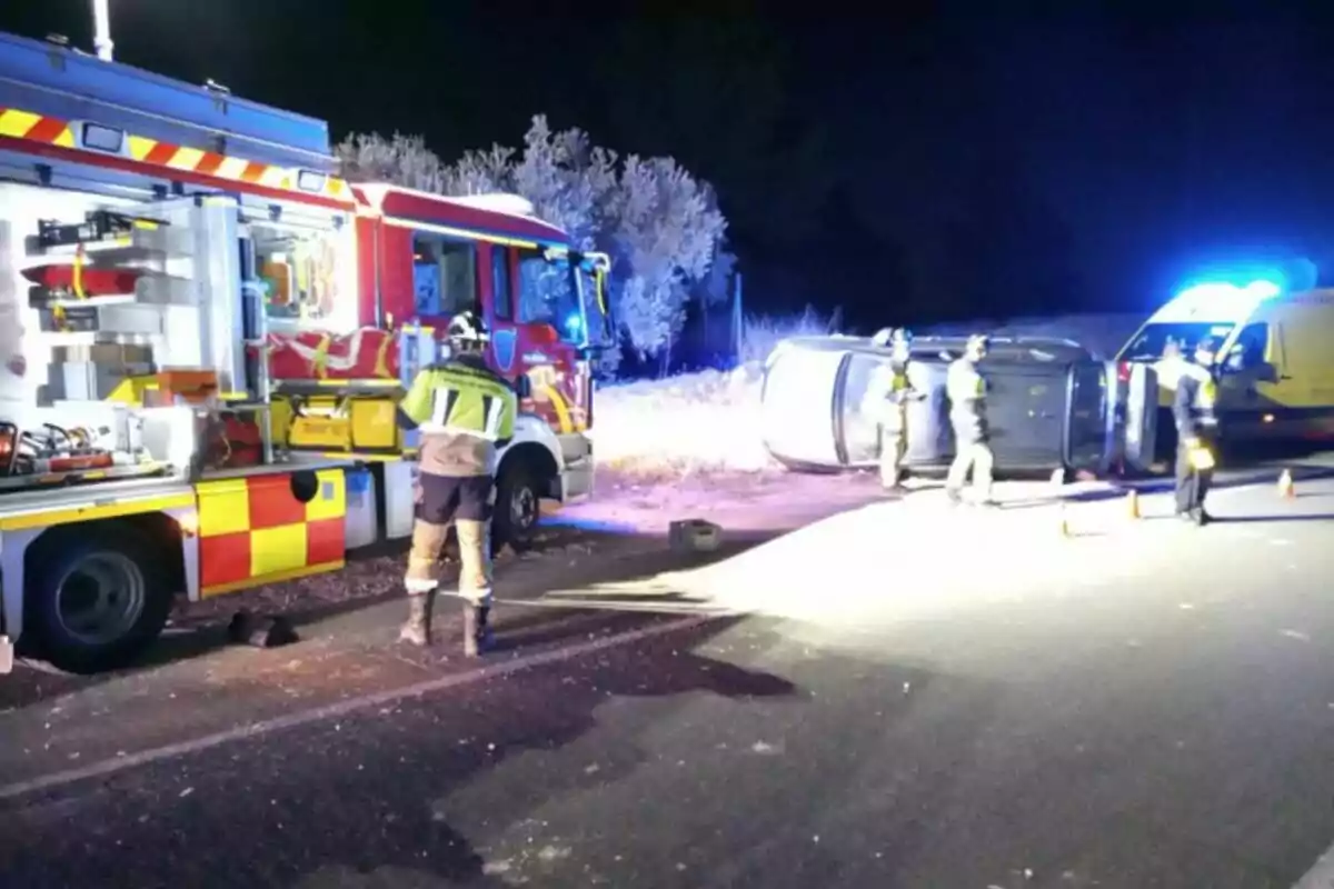 Un camión de bomberos y una ambulancia en la escena de un accidente de tráfico con un coche volcado en la carretera mientras los bomberos trabajan en el lugar.