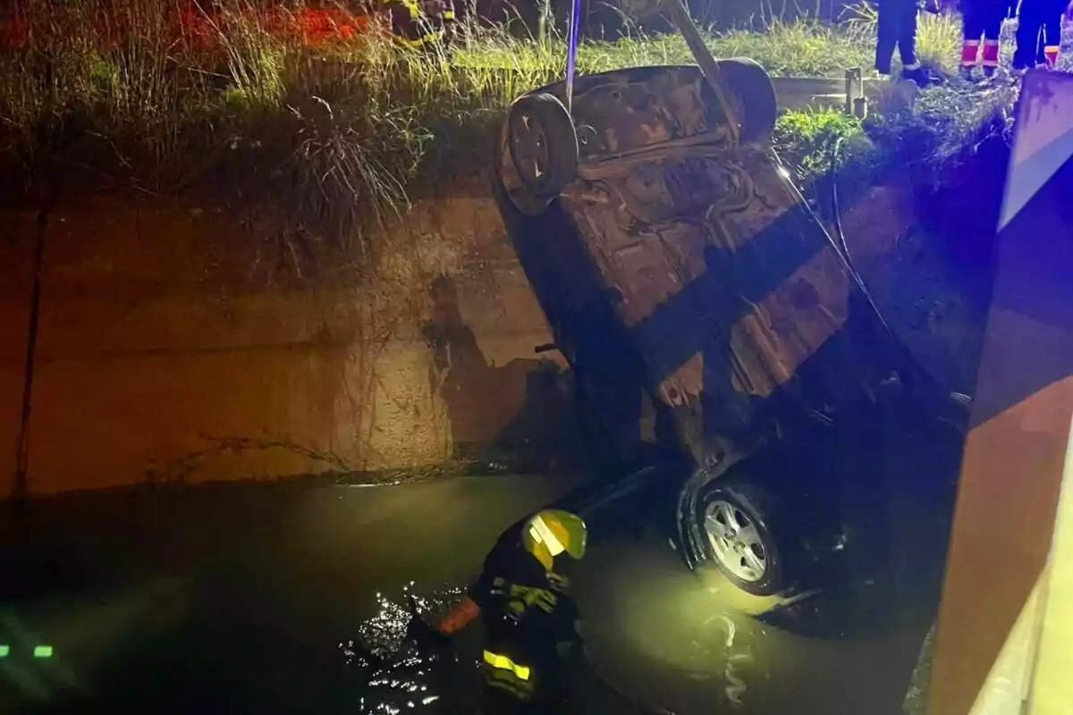 Un coche volcado es rescatado de un canal de agua por un bombero durante la noche.