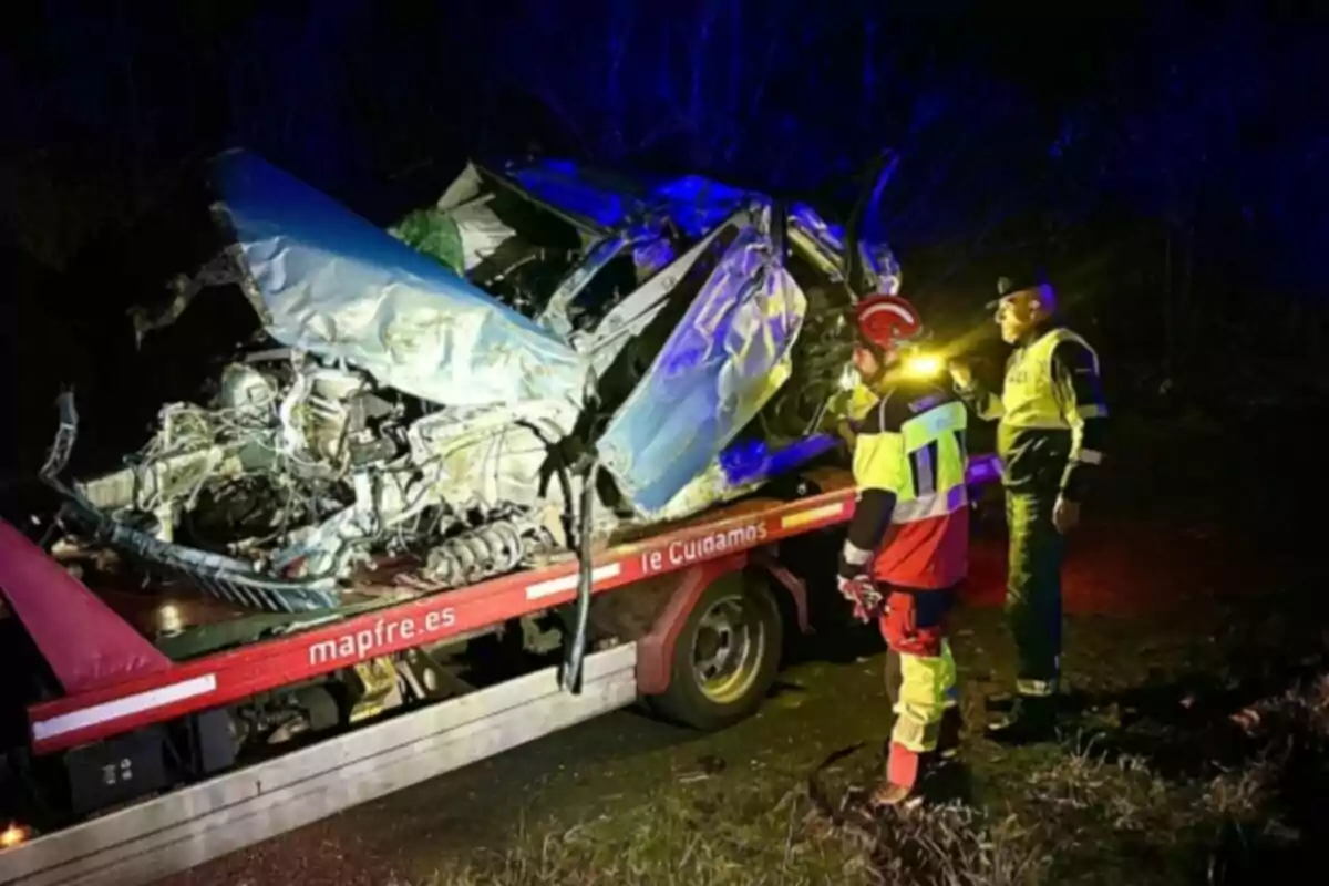 Dos personas con chalecos reflectantes observan un vehículo gravemente dañado sobre una grúa en una carretera oscura.