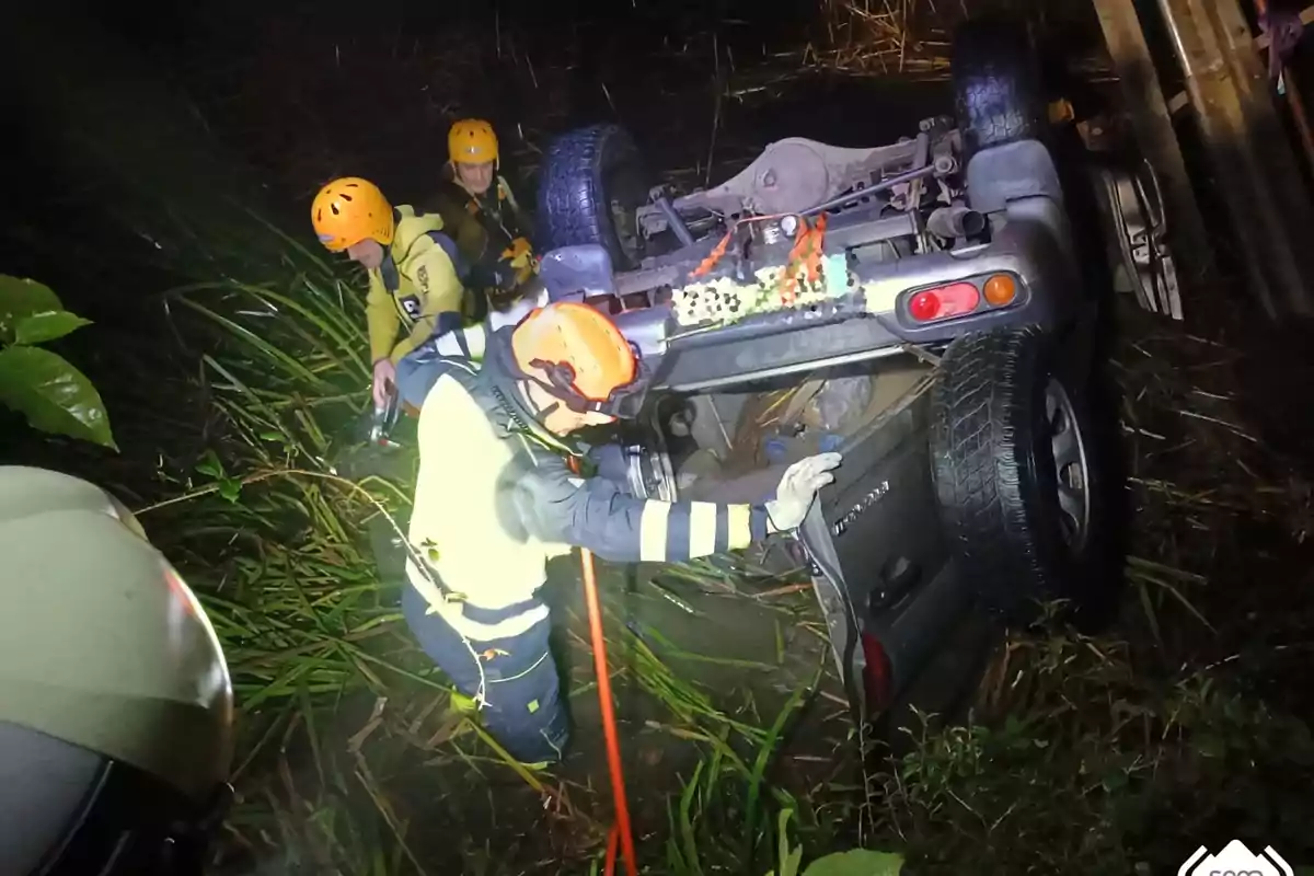 Rescatistas con cascos y equipo de seguridad trabajan en el lugar de un accidente donde un vehículo está volcado sobre su techo en un área con vegetación.