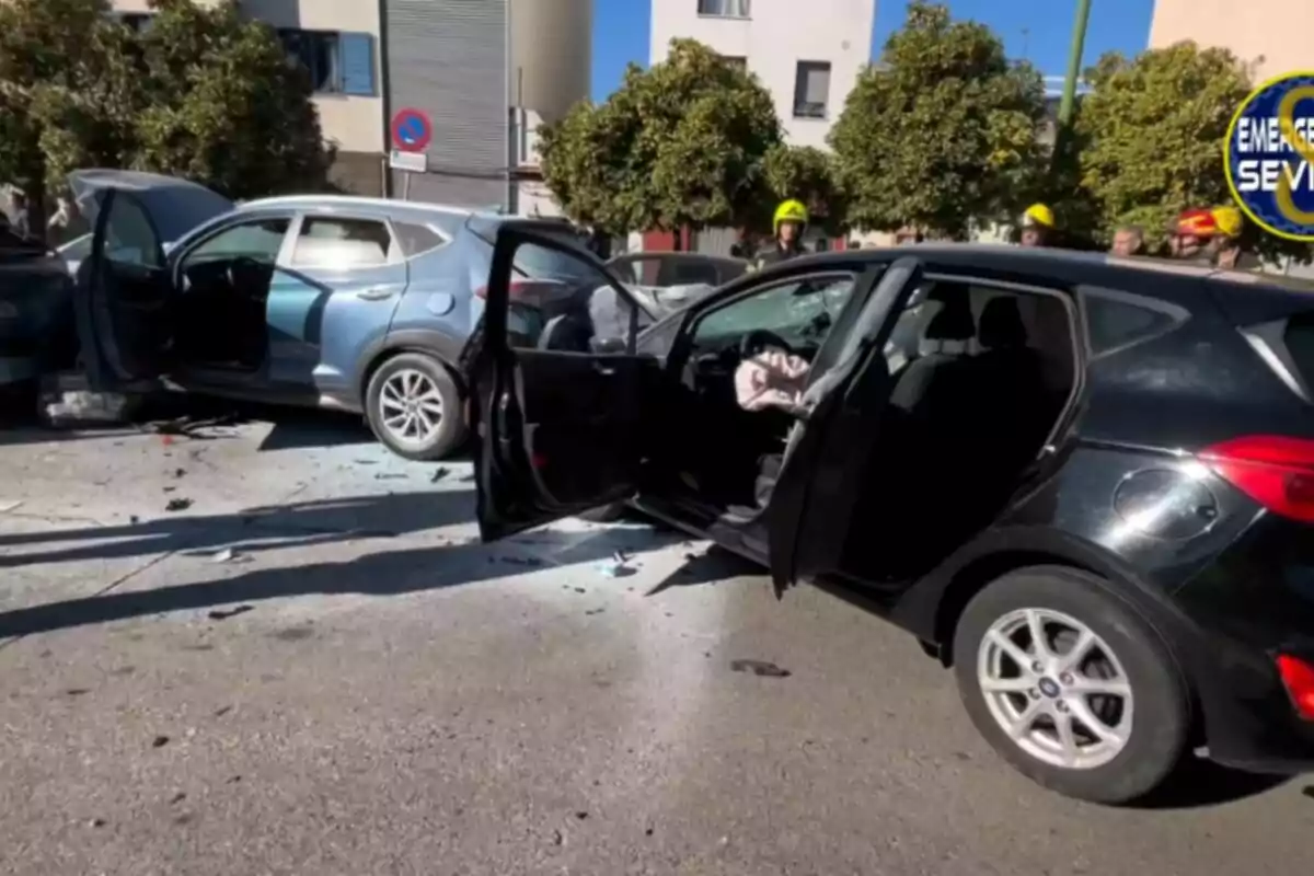 Dos coches involucrados en un accidente con las puertas abiertas y daños visibles en una calle urbana con árboles y edificios al fondo.