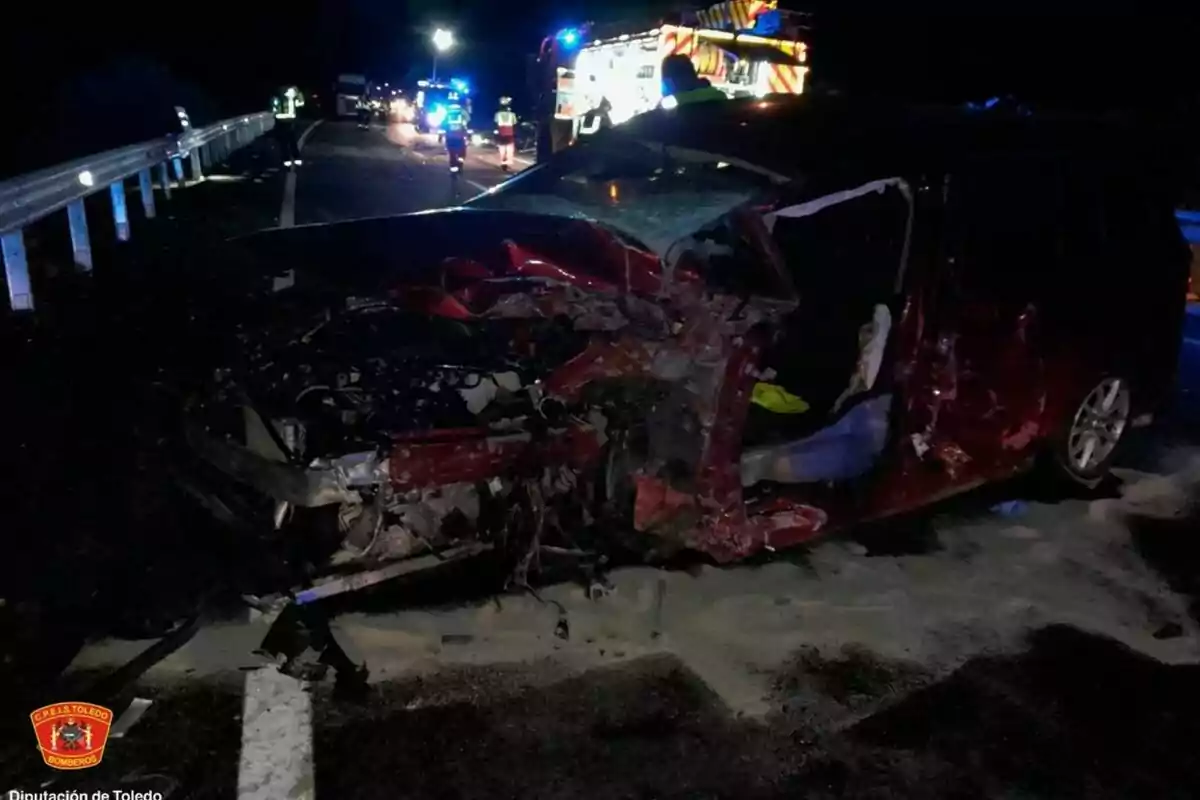 Un coche rojo gravemente dañado en un accidente nocturno en una carretera con presencia de bomberos y vehículos de emergencia iluminados al fondo.