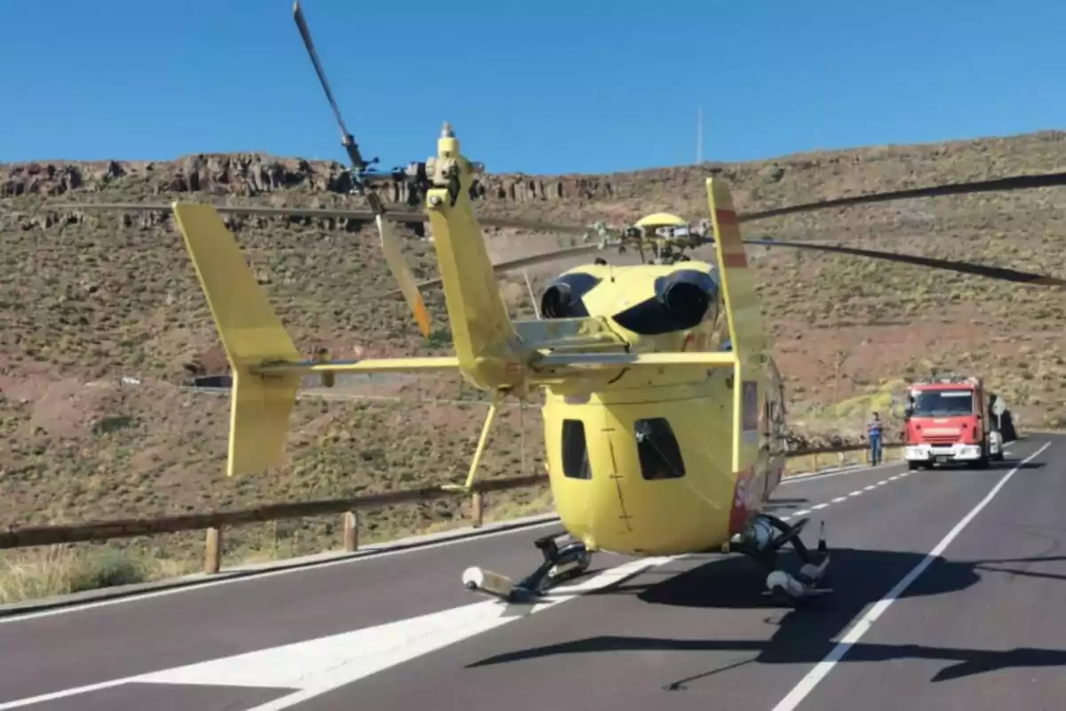Un helicóptero amarillo aterrizado en una carretera con un camión de bomberos al fondo y un paisaje montañoso. Un helicóptero amarillo aterrizado en una carretera con un camión de bomberos al fondo y un paisaje montañoso.