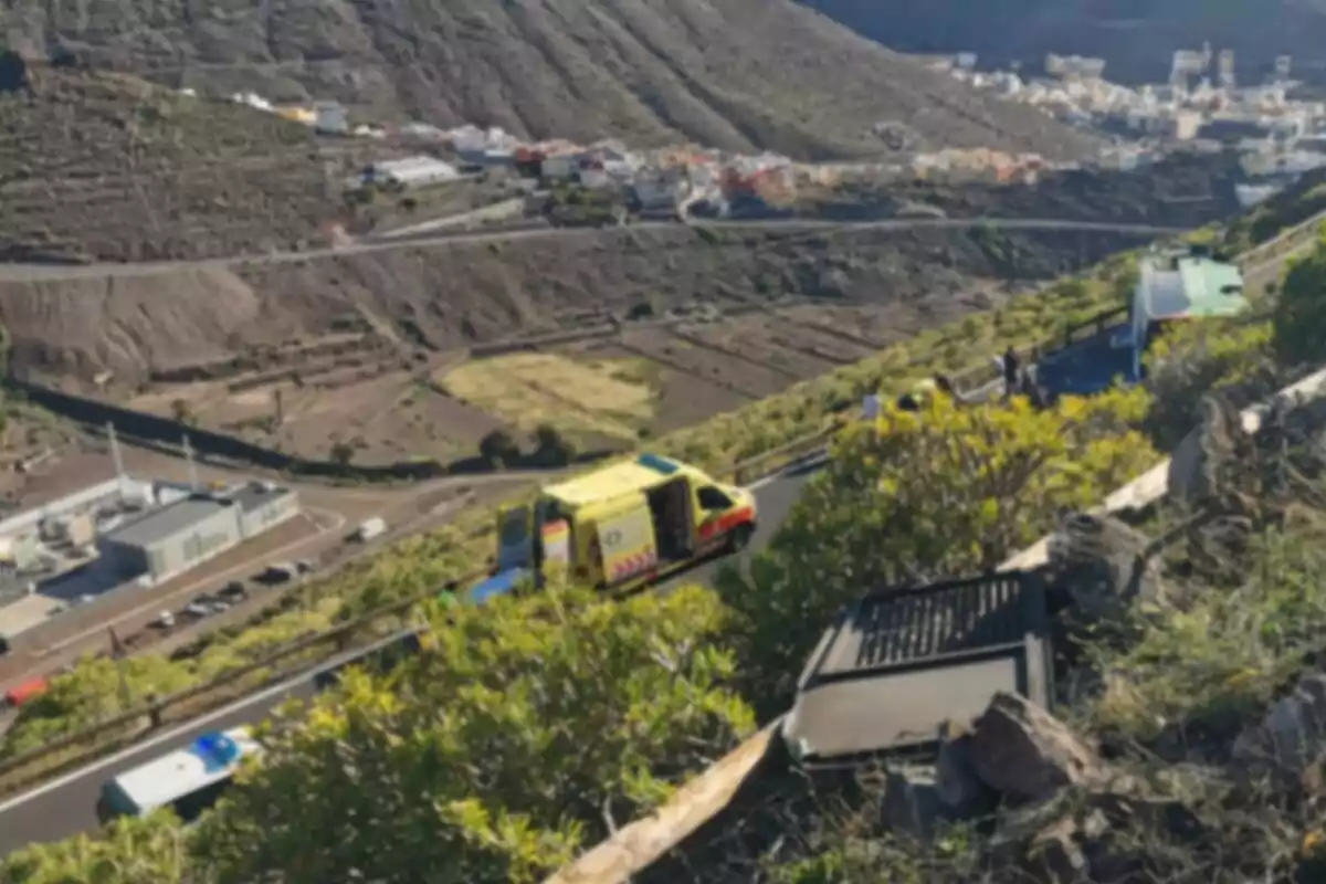 Una carretera de montaña con una ambulancia amarilla y varios vehículos en la vía, rodeada de vegetación y un paisaje montañoso al fondo. Una carretera de montaña con una ambulancia amarilla y varios vehículos en la vía, rodeada de vegetación y un paisaje montañoso al fondo.