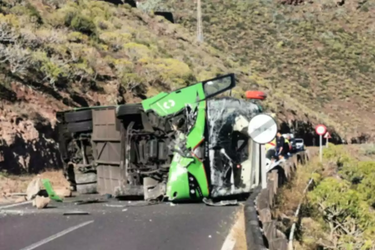 Un autobús verde volcado en una carretera de montaña con daños visibles en el vehículo y algunos coches y personas al fondo.