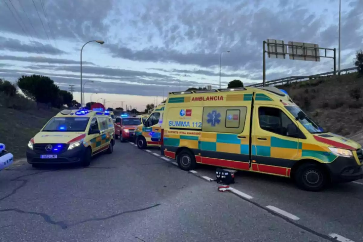 Varias ambulancias y vehículos de emergencia están estacionados en una carretera al atardecer con luces azules encendidas.