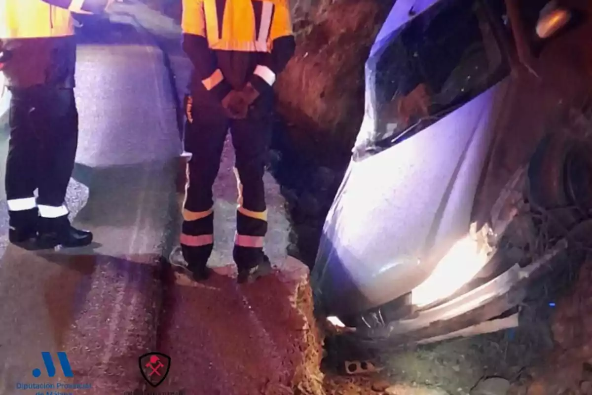 Un coche se encuentra parcialmente hundido en una zanja al lado de una carretera mientras dos personas con uniformes de seguridad observan la escena.