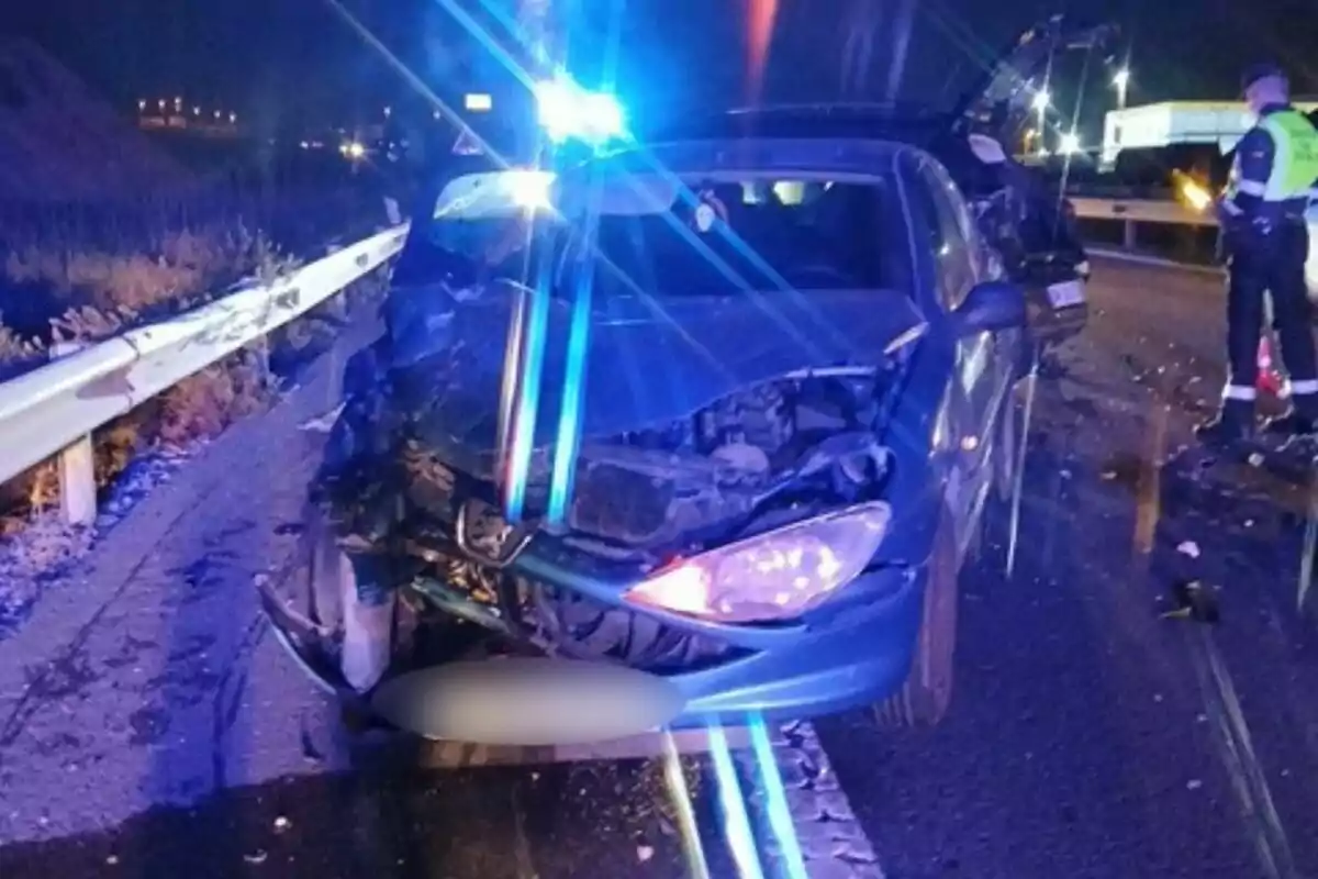 Un coche con el frente dañado tras un accidente en una carretera por la noche con luces de emergencia azules y un oficial de policía en la escena.