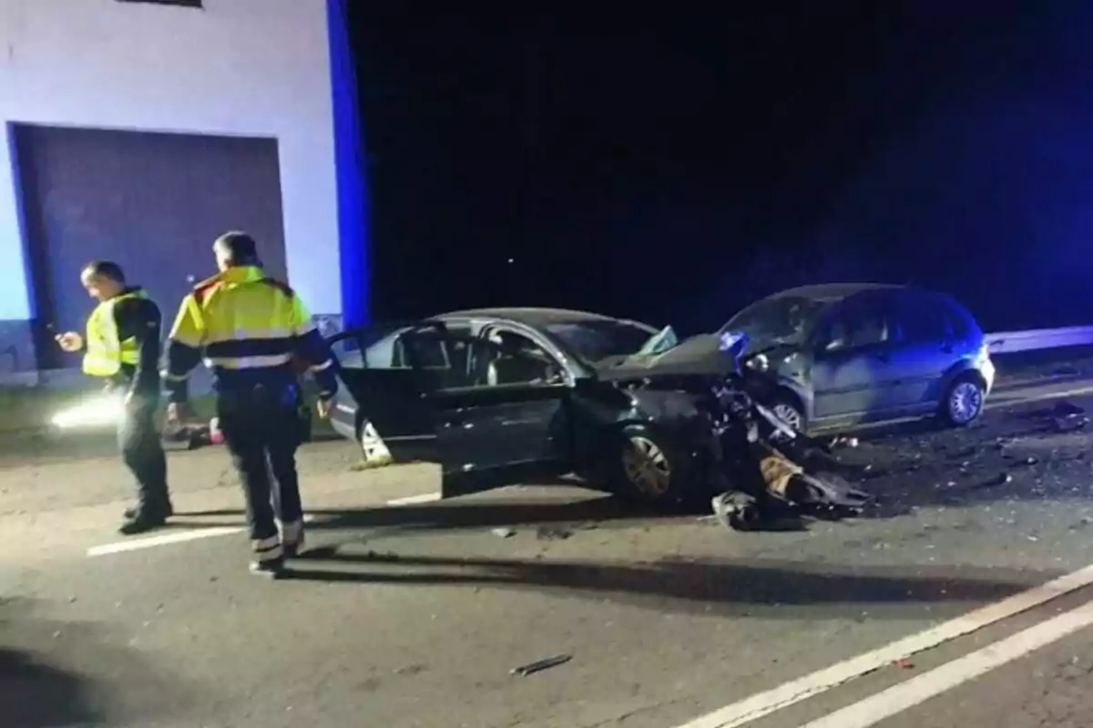 Accidente de tráfico nocturno con dos coches dañados y dos personas con chalecos reflectantes en la escena.