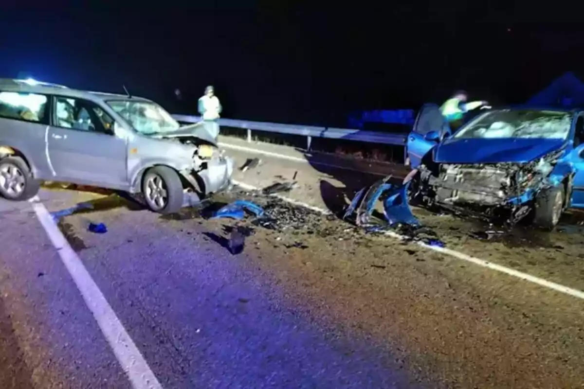 Dos coches chocados en una carretera por la noche con escombros esparcidos y personas observando.