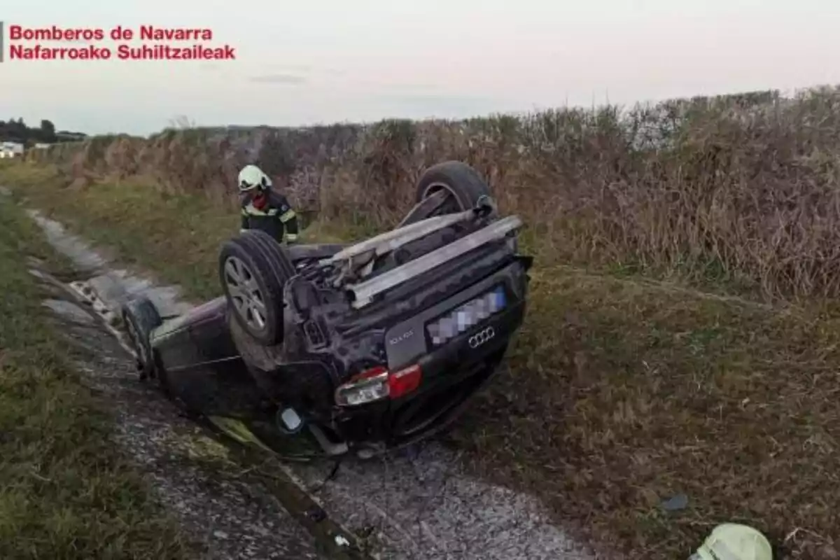 Un coche volcado en una zanja al lado de un camino rural con un bombero de pie cerca del vehículo.