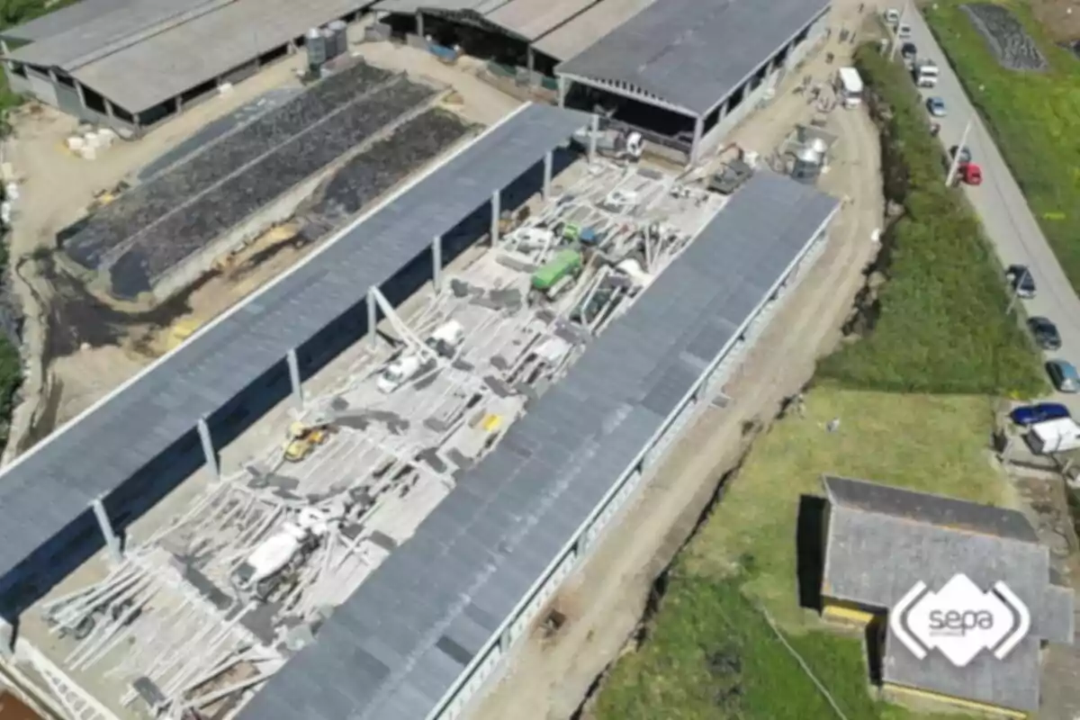 Vista aérea de una nave industrial en construcción con maquinaria y materiales en el interior rodeada de otras edificaciones y una carretera con varios vehículos estacionados Vista aérea de una nave industrial en construcción con maquinaria y materiales en el interior rodeada de otras edificaciones y una carretera con varios vehículos estacionados