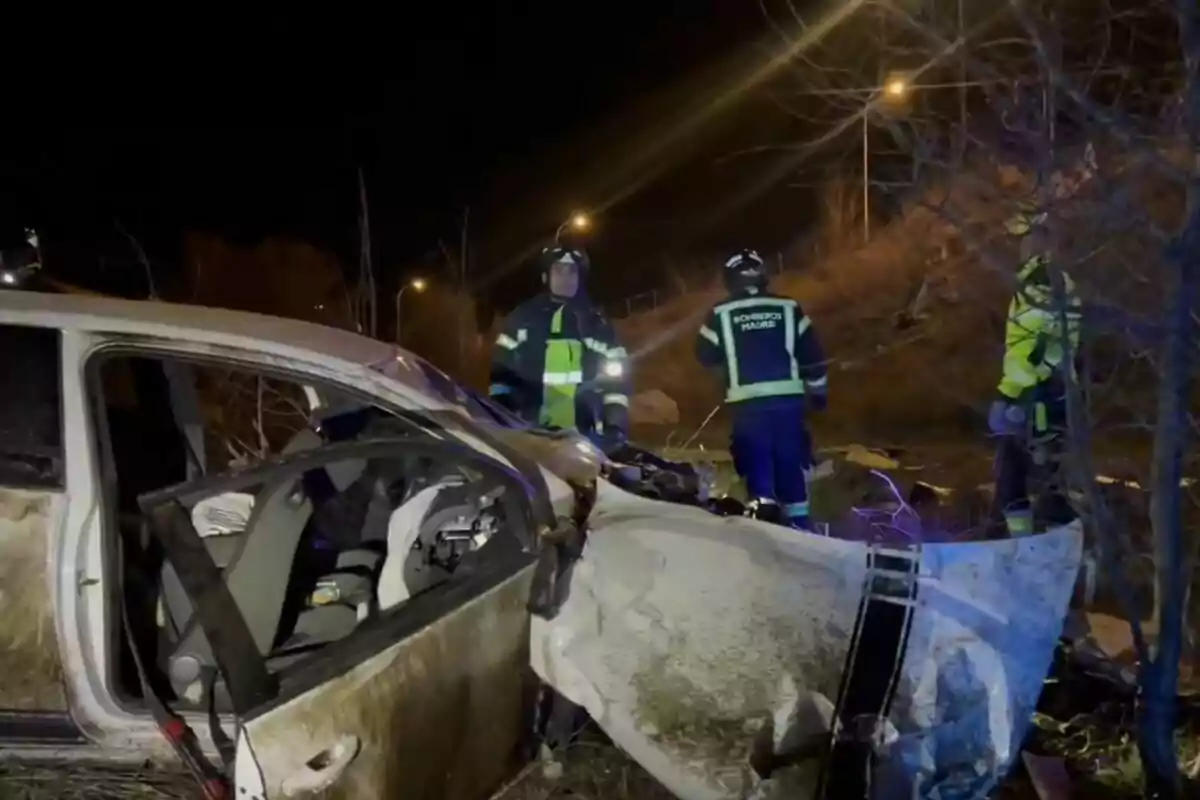 Un coche accidentado en la noche con bomberos trabajando en el lugar.